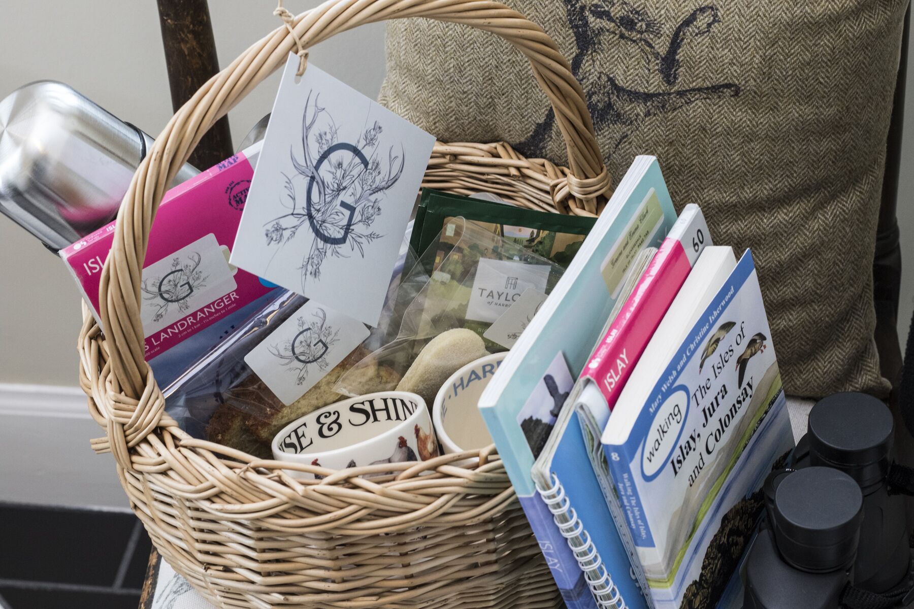Una cesta de mimbre llena de libros, una taza blanca y una caja rosa.
