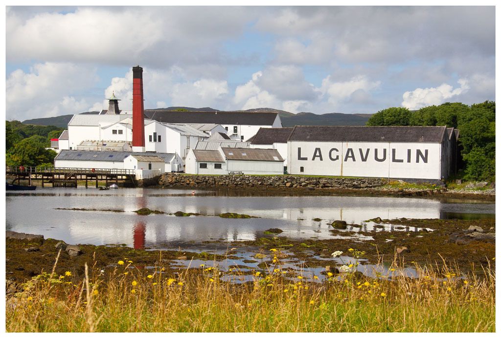 A historic building with a prominent chimney, named Lacavulin, situated by a body of water with a dock.