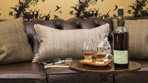 A bottle of Lagavulin whisky, two glasses, and a coaster on a round table in front of a sofa with a tree and bird wallpaper. - Glenegedale House (Islay Island)