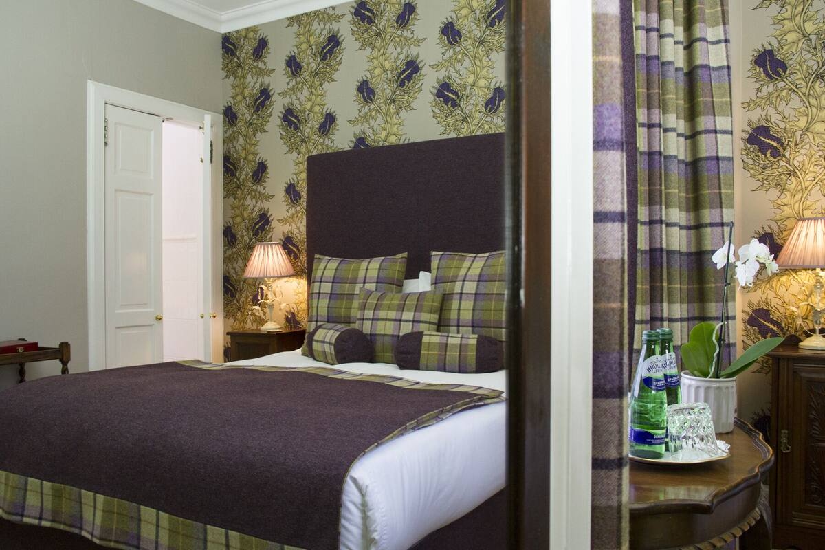A neatly made bed with a dark headboard, a bedside table with a lamp, and a floral wallpaper pattern.