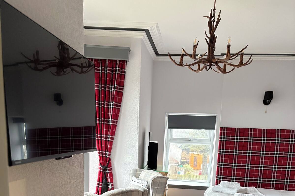A hotel room with a bed, a desk with a kettle, chairs, a TV, and a window with red curtains.