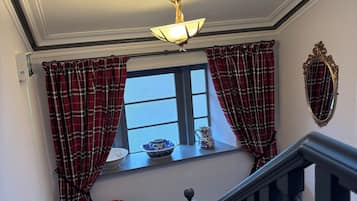 Een trap met houten leuning, een raam met geruite gordijnen, een decoratieve spiegel en een plafondlamp.