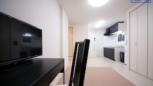A modern living room with a flat-screen TV on a dark wooden table, a black chair, and a brown rug. A kitchen with white cabinets and a black range hood is visible in the background. - Apartment Heights HANA (Sakai)