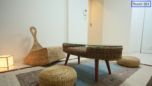 A room with a glass-top table, woven poufs, and a patterned rug. - Apartment Heights HANA (Sakai)