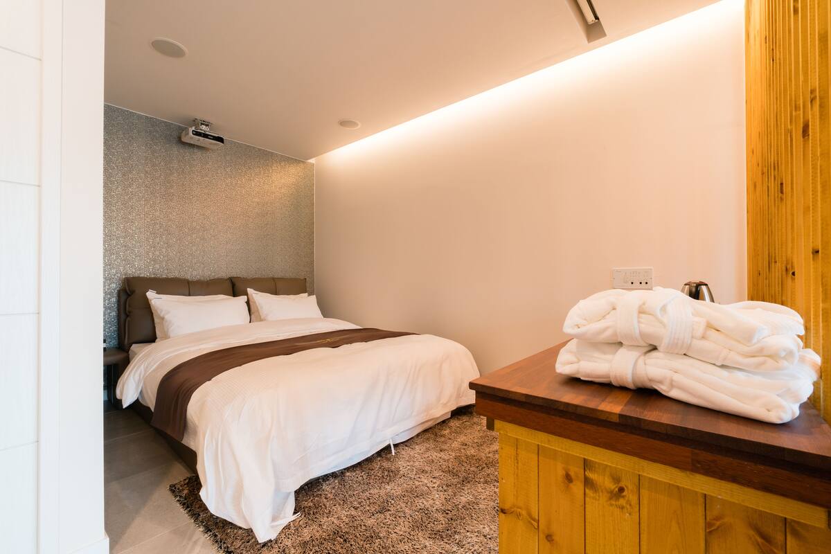 Ein Schlafzimmer mit Bett, einer hölzernen Sitzbank mit Handtüchern, einer strukturierten Wand und einer Deckenleuchte.