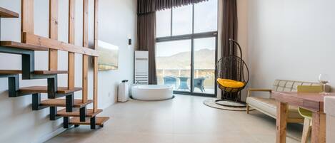 A modern living room with a wooden staircase, a round bathtub, a dining table, and a view of the outdoors.