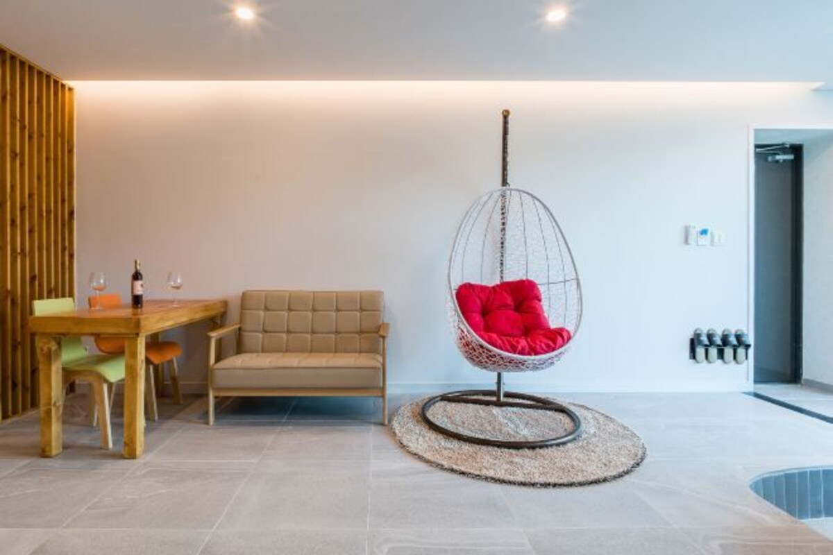 A modern living room with a wooden dining table, a beige sofa, and a red hanging chair.