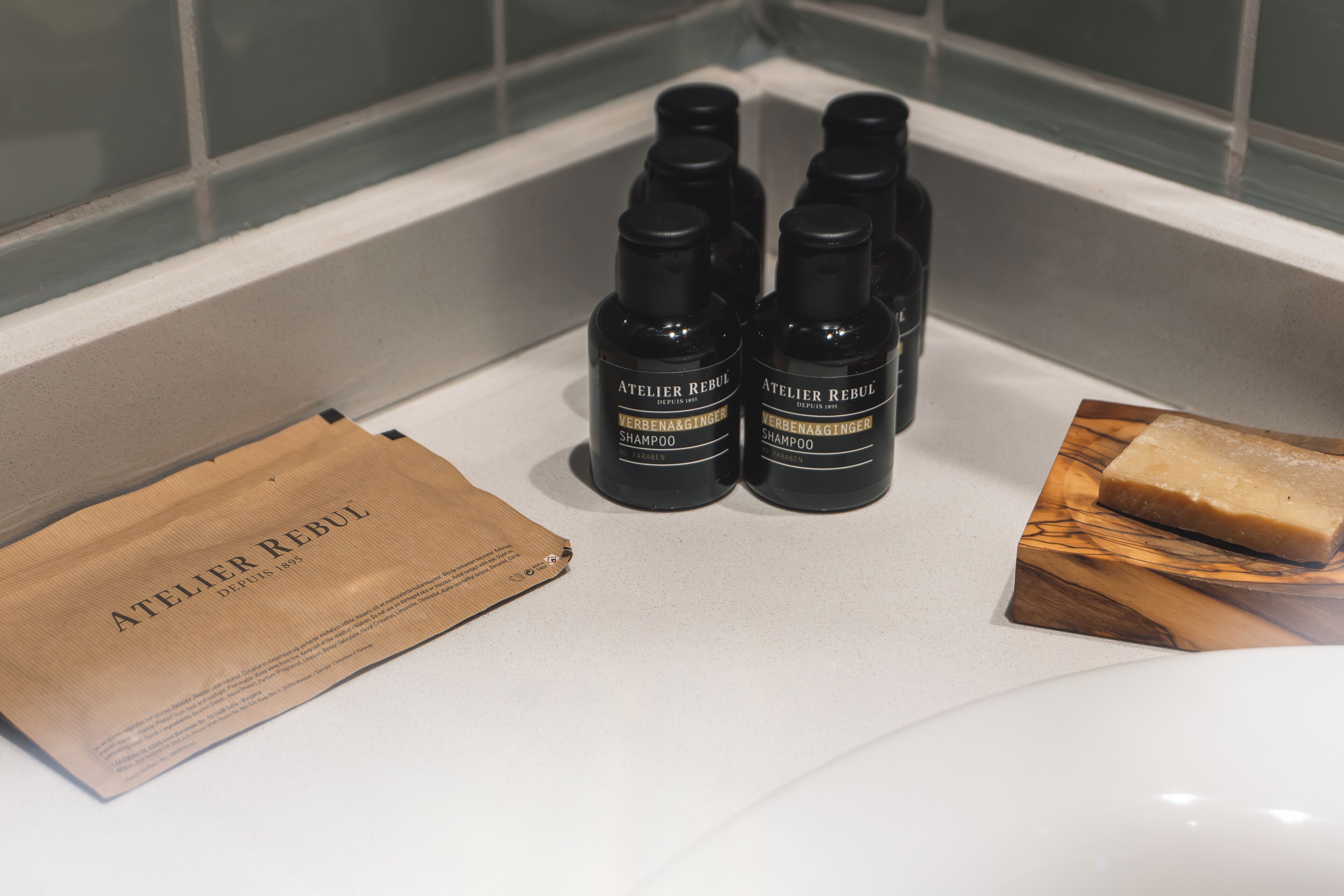 A bathroom with two bottles of Atelier Rebul shampoo, a wooden soap dish, and a brown paper bag.