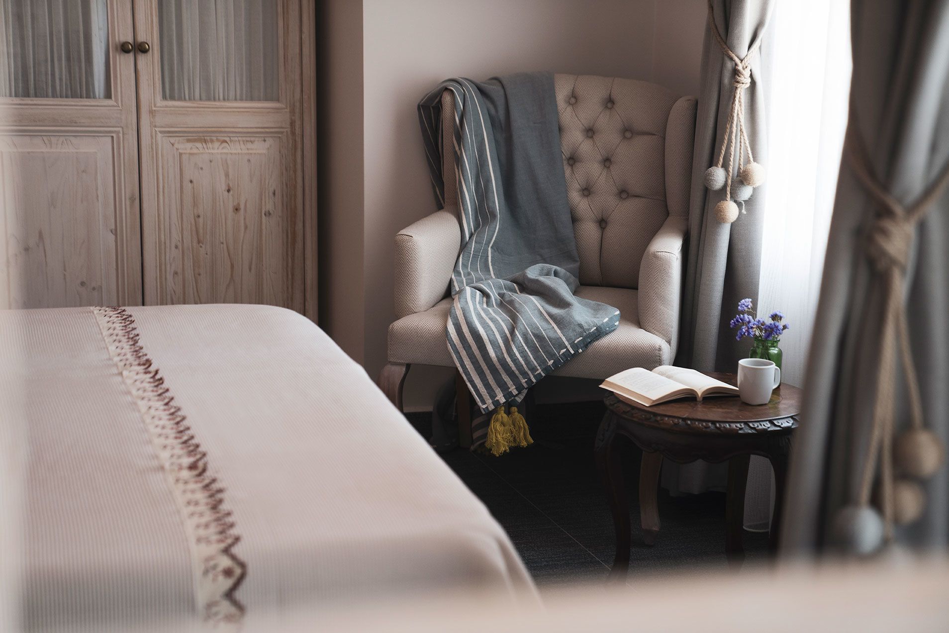 A neatly arranged bedroom with a bed, a chair with a striped blanket, a small table with a book and a mug, and a window with curtains.