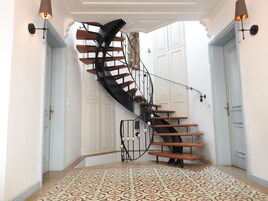 Uma escadaria em espiral com degraus de madeira e grades de metal preto, que leva a um patamar com ladrilhos estampados.