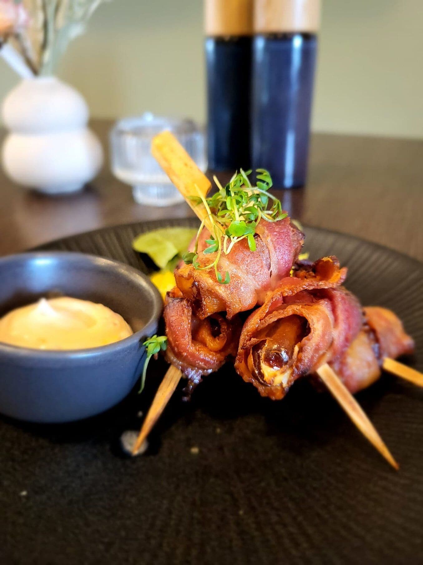 A plate with bacon-wrapped meat skewers, a small bowl of sauce, and a decorative garnish.