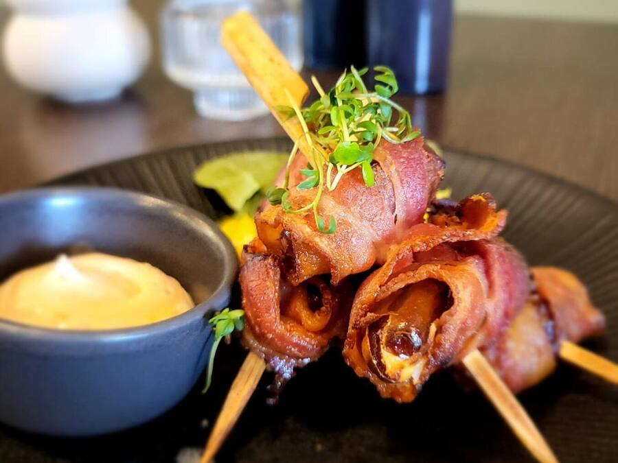 A plate with bacon-wrapped meat skewers, a small bowl of sauce, and a decorative garnish.