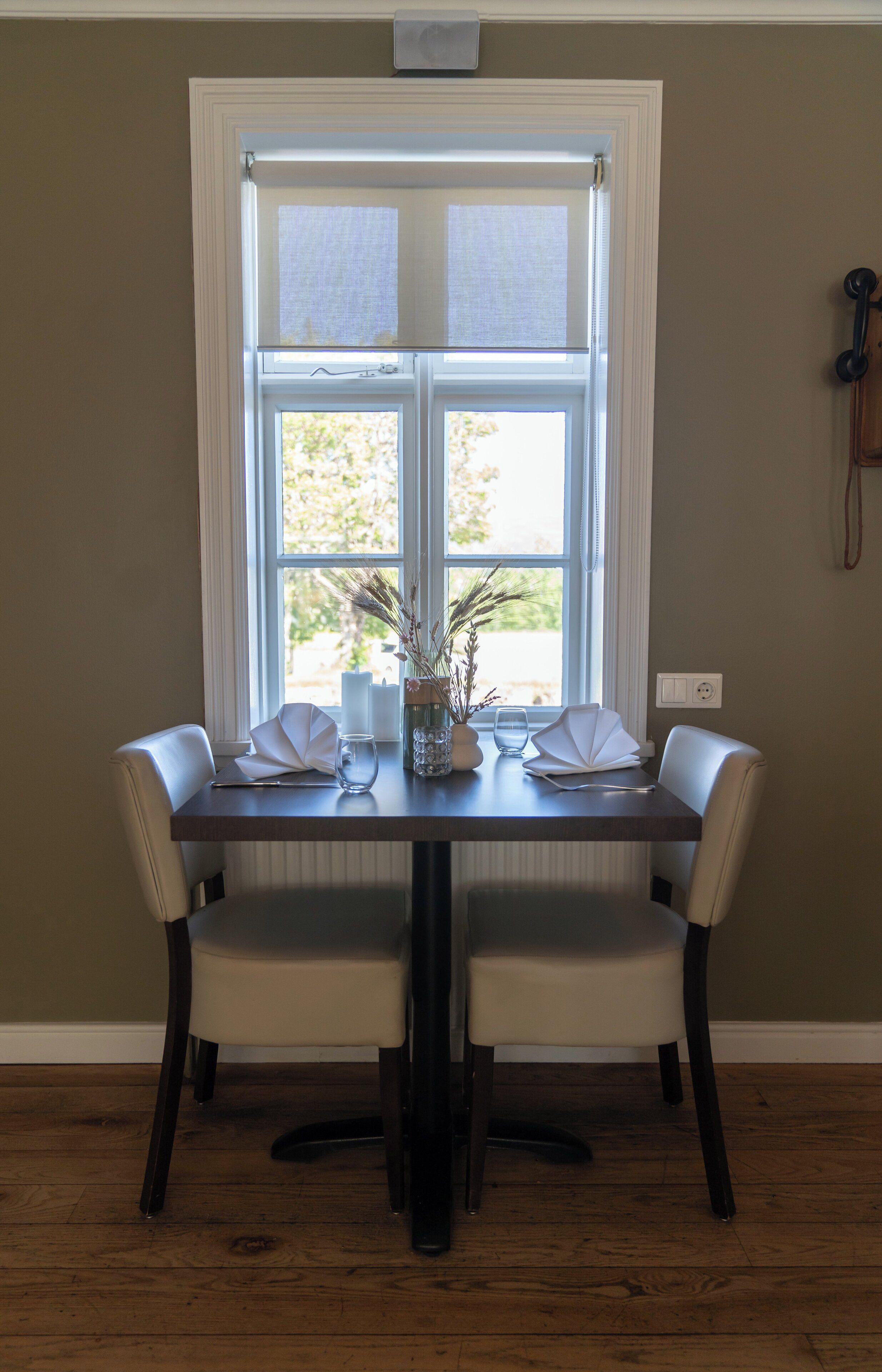 A dining table with two chairs, a vase with dried plants, and a window with curtains.