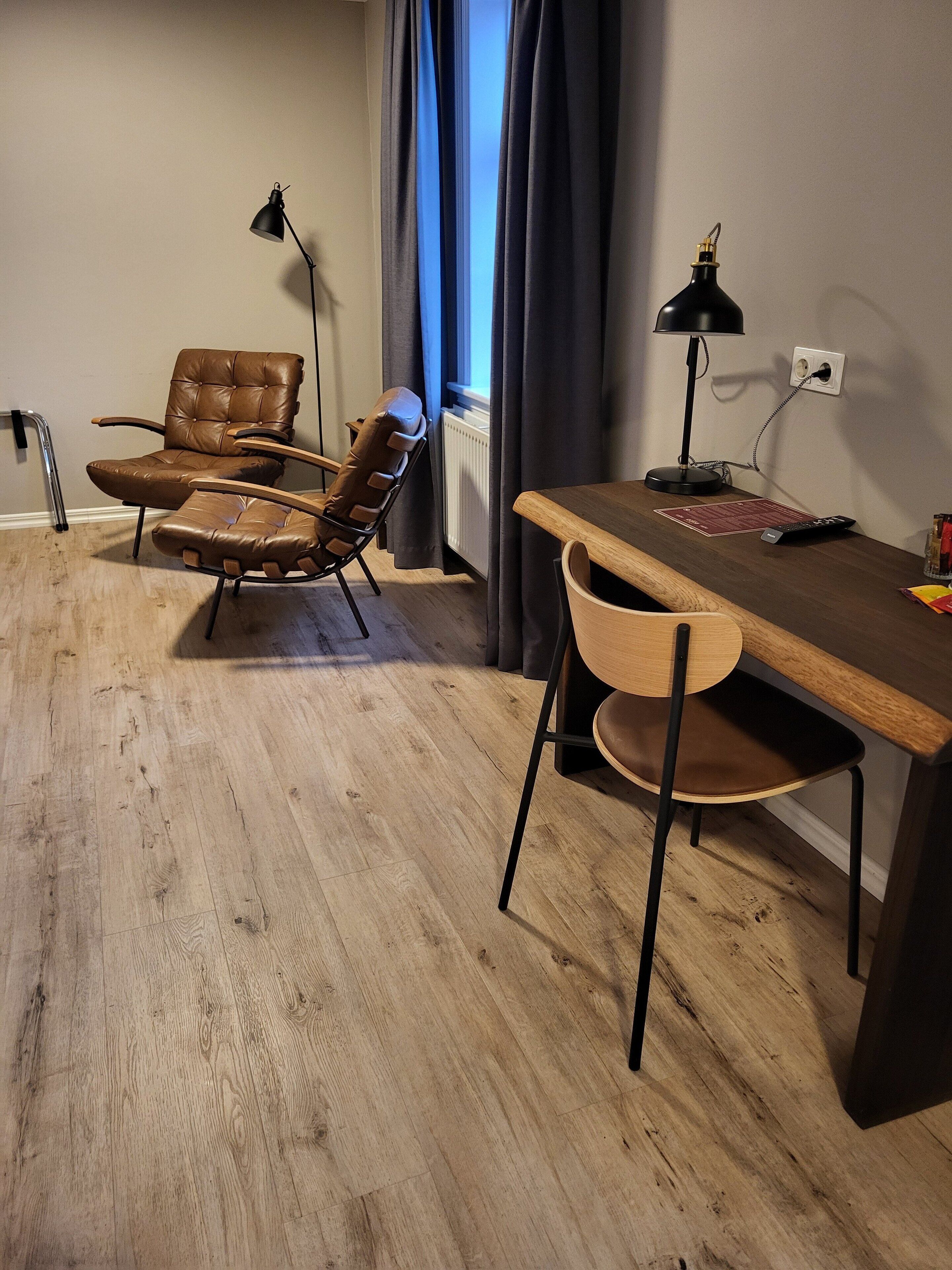 Una habitación con piso de madera, un escritorio con lámpara, dos sillones de cuero, un radiador y una ventana con cortinas.