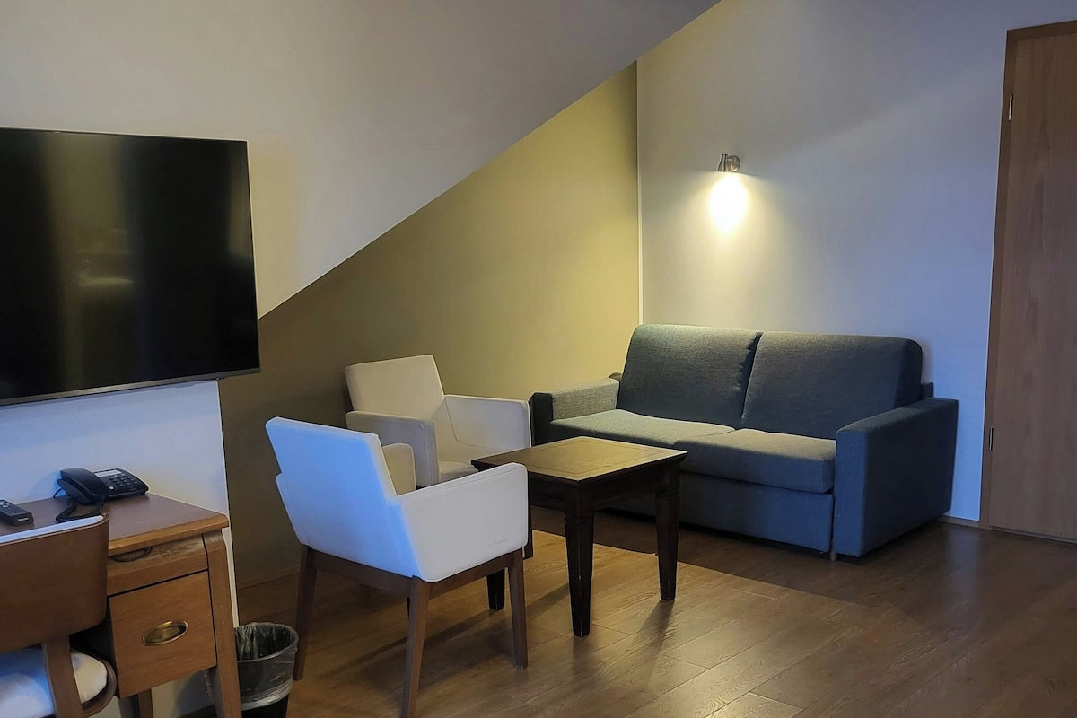 Una sala de estar moderna con piso de madera, un sofá gris, un sillón blanco, una mesa de centro de madera y un televisor de pantalla plana montado en la pared.