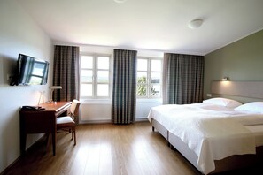 A hotel room with a bed, a desk with a chair, a television mounted on the wall, and a window with curtains.