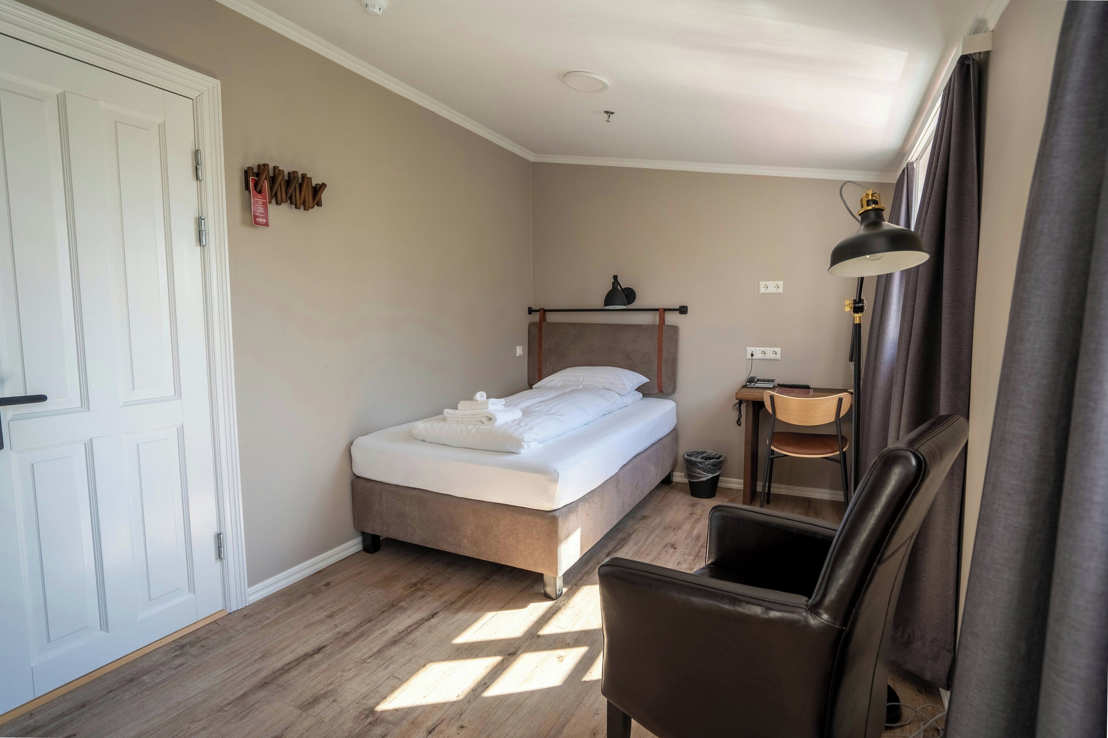 Ein Schlafzimmer mit einem Bett, einem Schreibtisch, einem Stuhl, einer Lampe und einem Fenster mit Vorhängen.