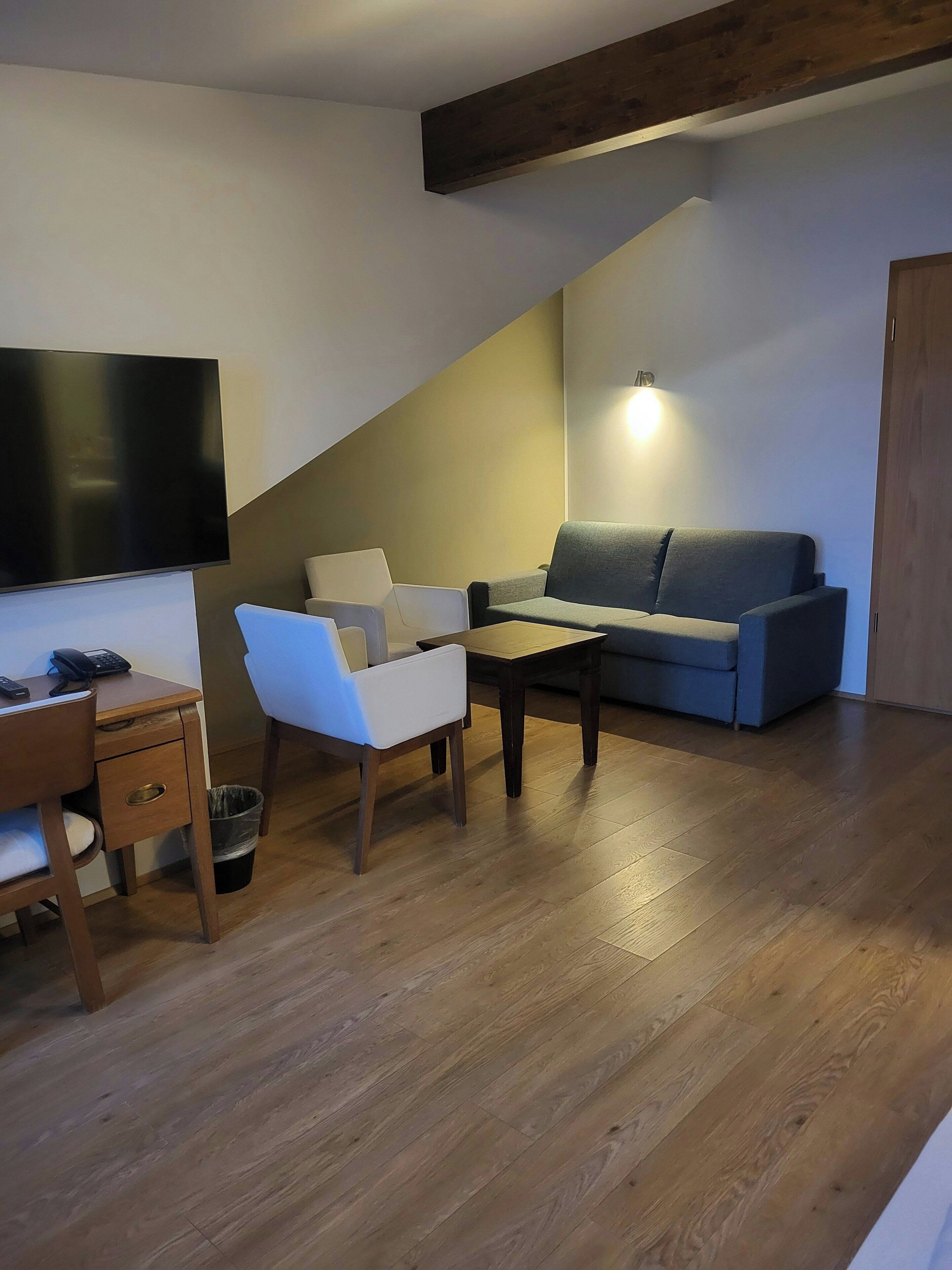 A modern living room with a wooden floor, a grey sofa, a white armchair, a wooden coffee table, and a flat-screen TV mounted on the wall.