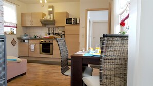 A compact kitchen with a microwave, oven, and various utensils. There is a dining area with a table set for two, and a wicker chair.