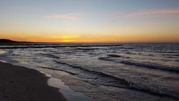 A beach with waves at sunset, a pier extending into the water, and a distant shoreline.