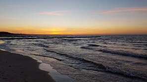 A beach with waves at sunset, a pier extending into the water, and a distant shoreline.