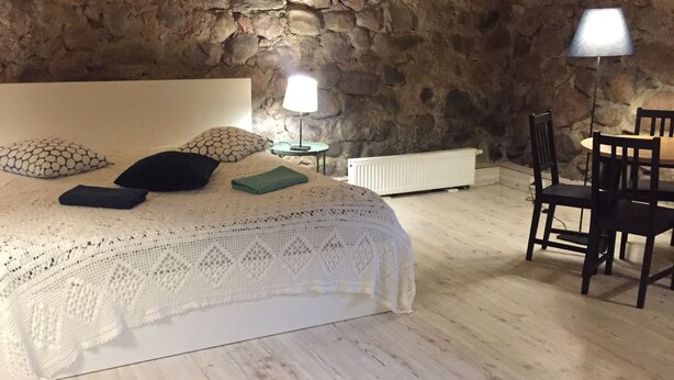 A bedroom with a stone wall, a bed with a white knitted blanket, two bedside lamps, a radiator, and a wooden dining table with chairs. - Sleep In BnB (Vilnius)