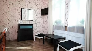 A room with a wooden floor, a black desk, a white bench, a black cabinet, a framed picture on the wall, and a window with sheer curtains. - Sleep In BnB (Vilnius)