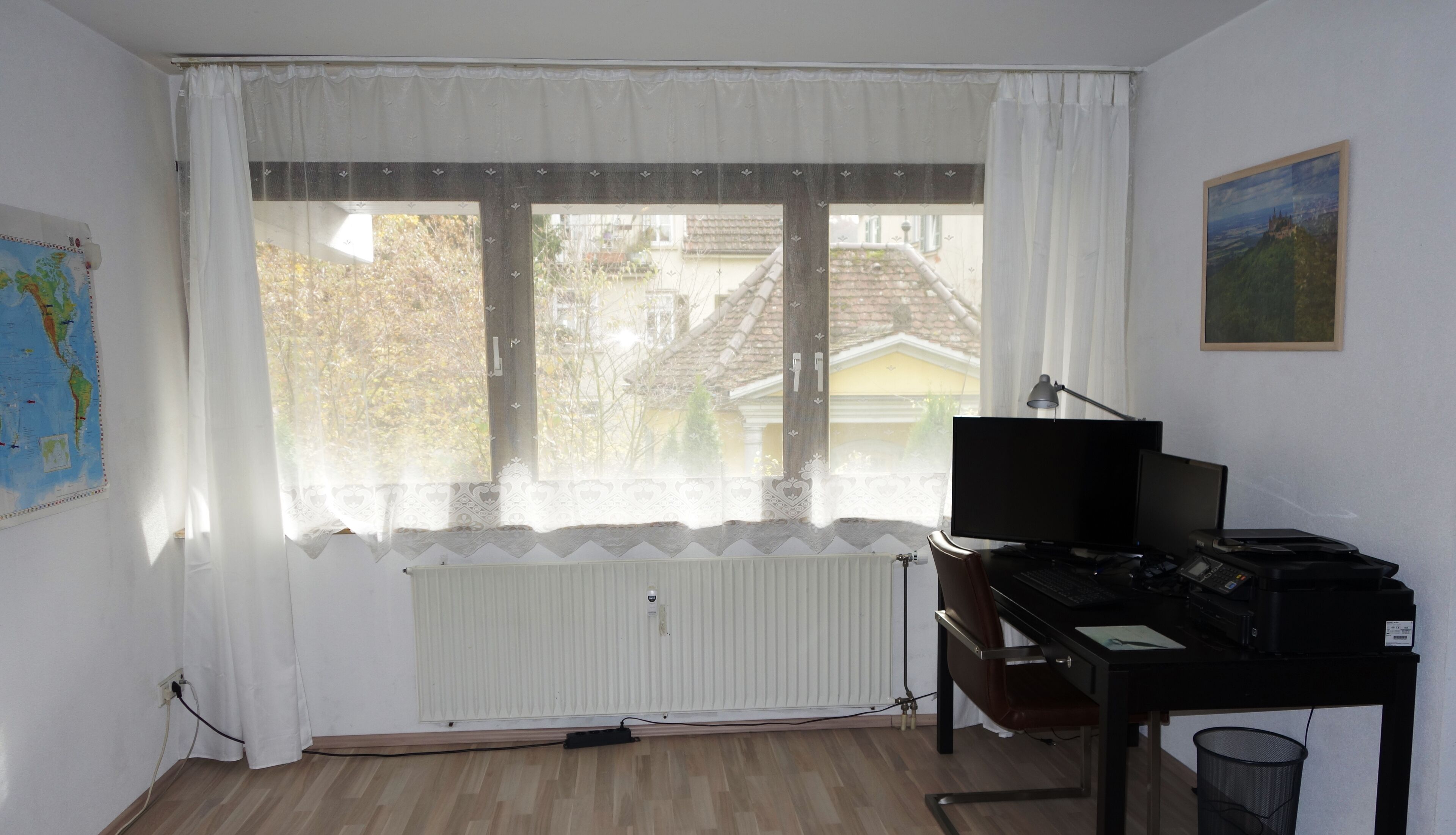 A room with a large window, a desk with a computer, a chair, a radiator, and a map on the wall.