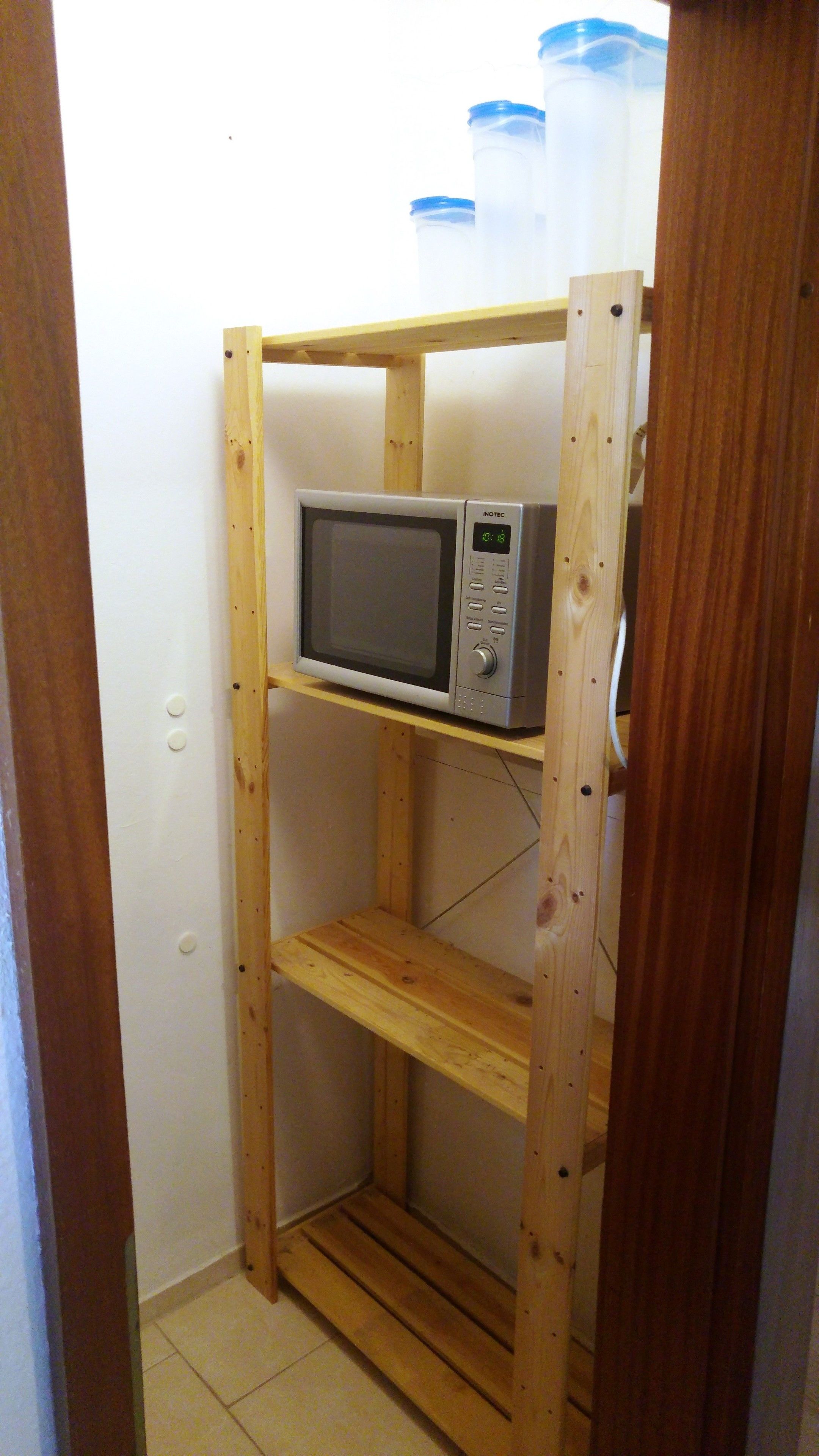 Une étagère en bois avec un micro-ondes sur l’étagère supérieure, une prise de courant sur le mur et un cadre de porte en bois.