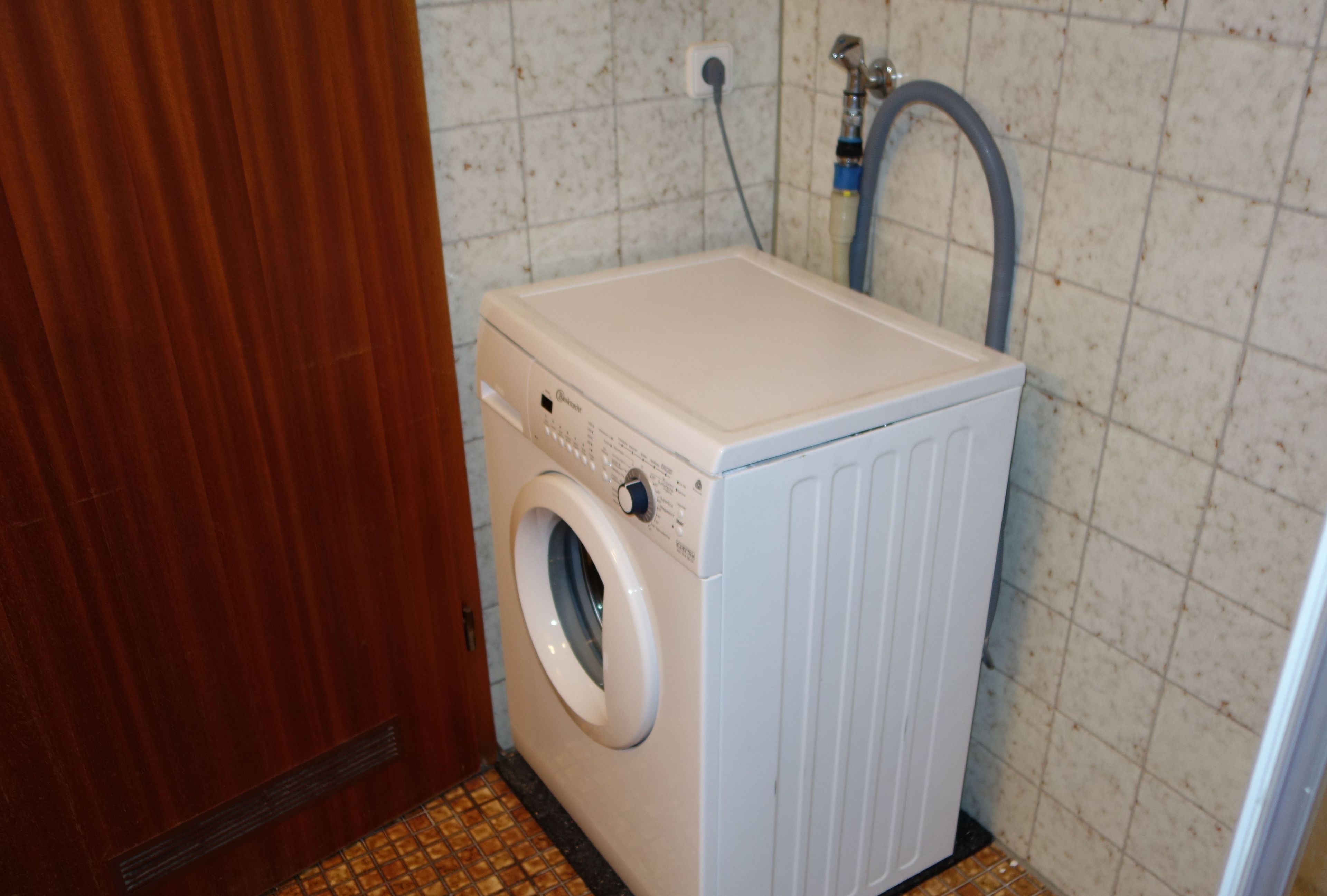 Une petite machine à laver blanche, dans une salle de bain de taille réduite, dont les murs sont recouverts de carreaux et qui possède une porte en bois.