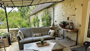 A cozy outdoor seating area with a wicker sofa, a wooden table, and a stone wall backdrop. - Le Vallonnet gîte (Le Vigan)
