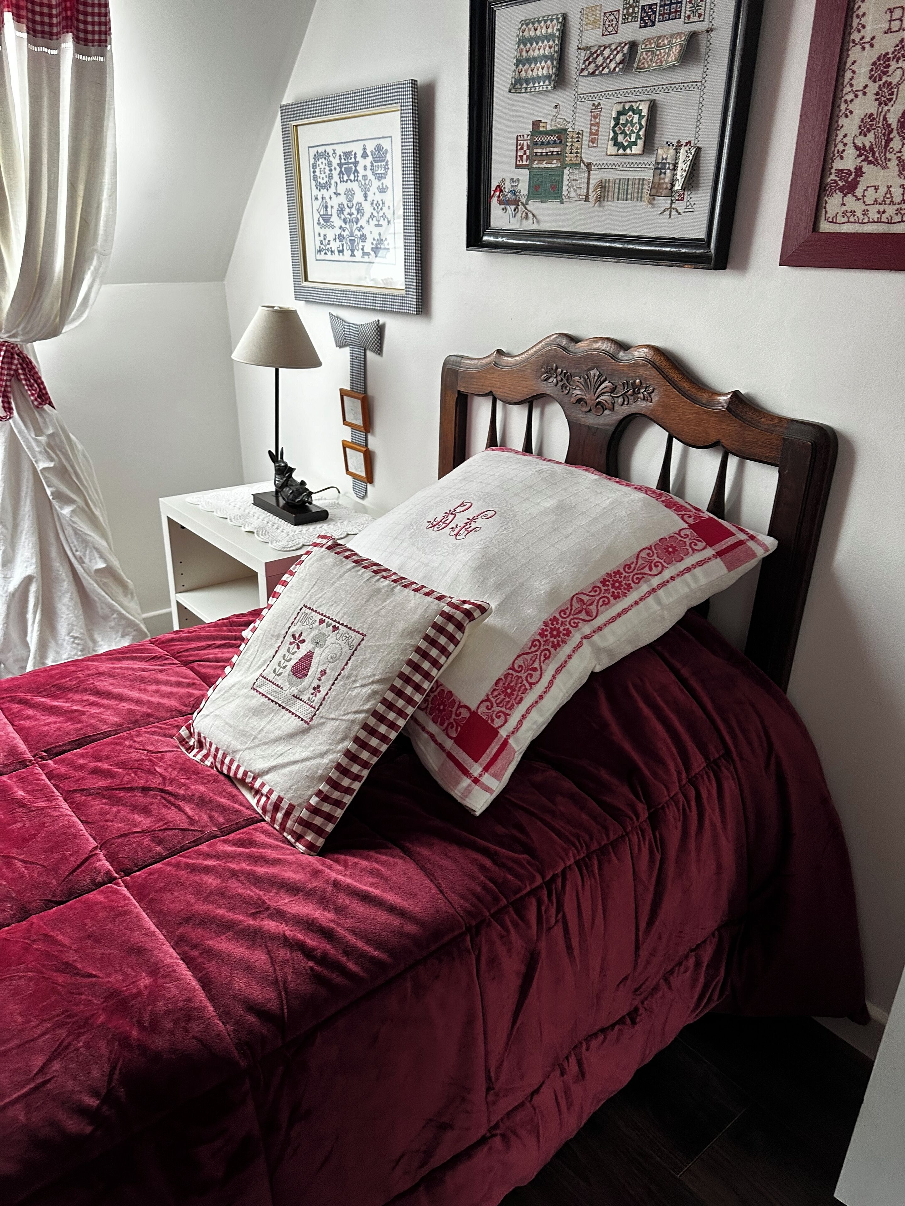 Un dormitorio con cabecera de madera, colcha acolchada roja y almohada blanca con bordado. Encima de la cama hay una obra de arte textil enmarco y una mesita de noche con una lámpara.