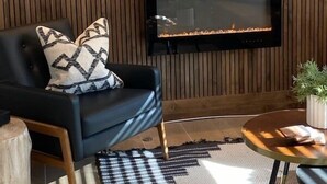 Una sala moderna con un revestimiento de madera, un sofá negro con cojines estampados, un televisor de pantalla plana sobre una chimenea y una mesa de centro redonda con una planta.