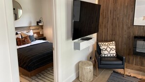 Un dormitorio moderno con una cama, un televisor montado en la pared, un panel de madera decorativo y una silla con un cojín estampado.
