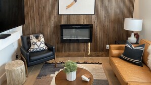Una sala moderna con un televisor de pantalla plana, un panel de pared de madera, un sofá de cuero negro, un sillón de cuero marrón y una mesa de centro de madera con una planta.