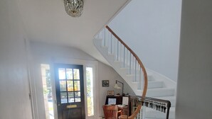 Una habitación amplia con suelo de madera, un escritorio con silla, una escalera con barandilla y una puerta que da al exterior.