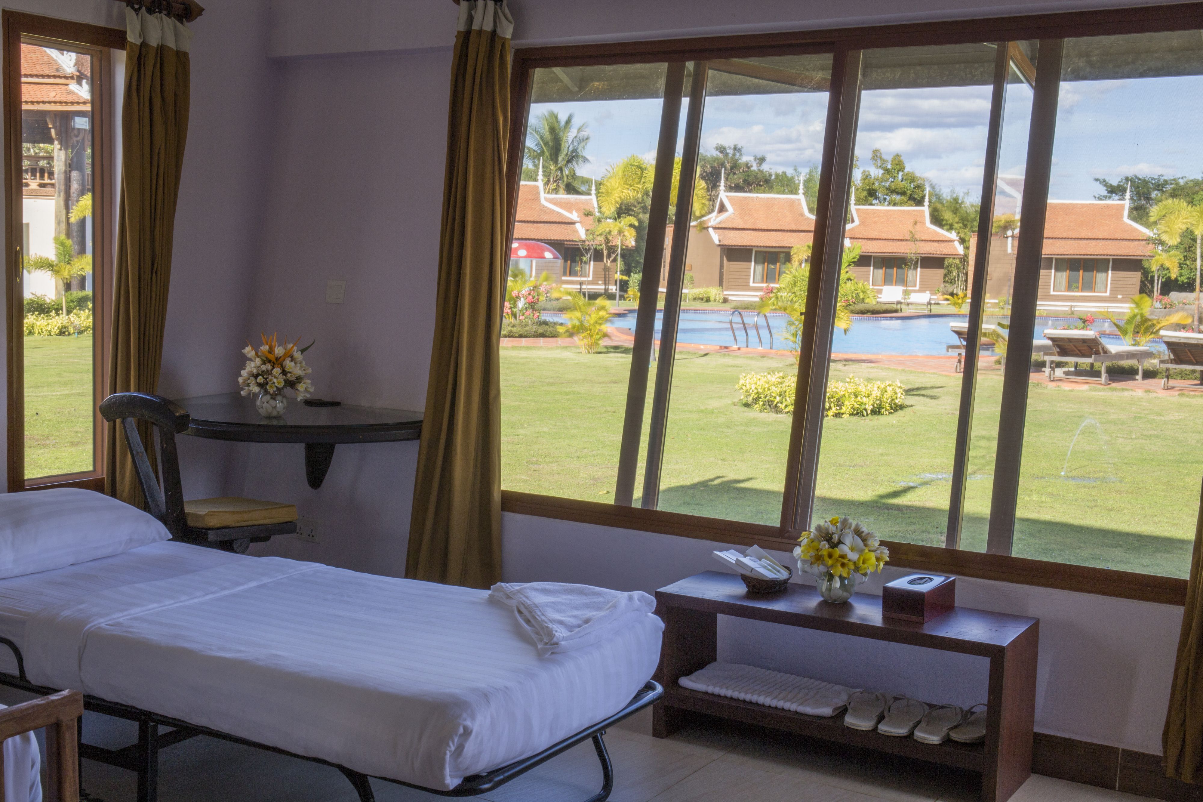 A hotel room with a bed, a desk with a telephone and flowers, and a view of a swimming pool and buildings.