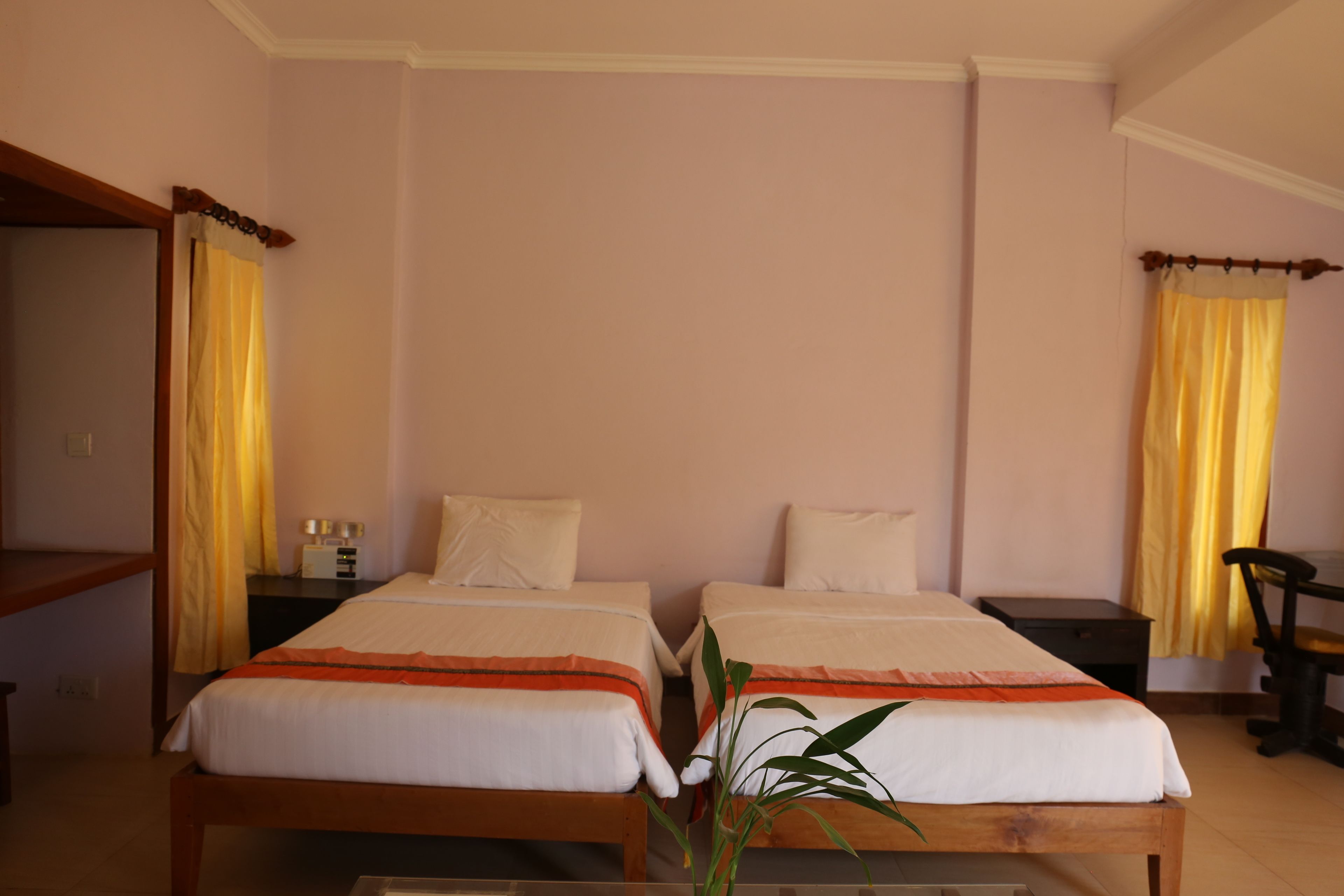 A room with two beds, wooden headboards, a bedside table with a lamp, and yellow curtains.