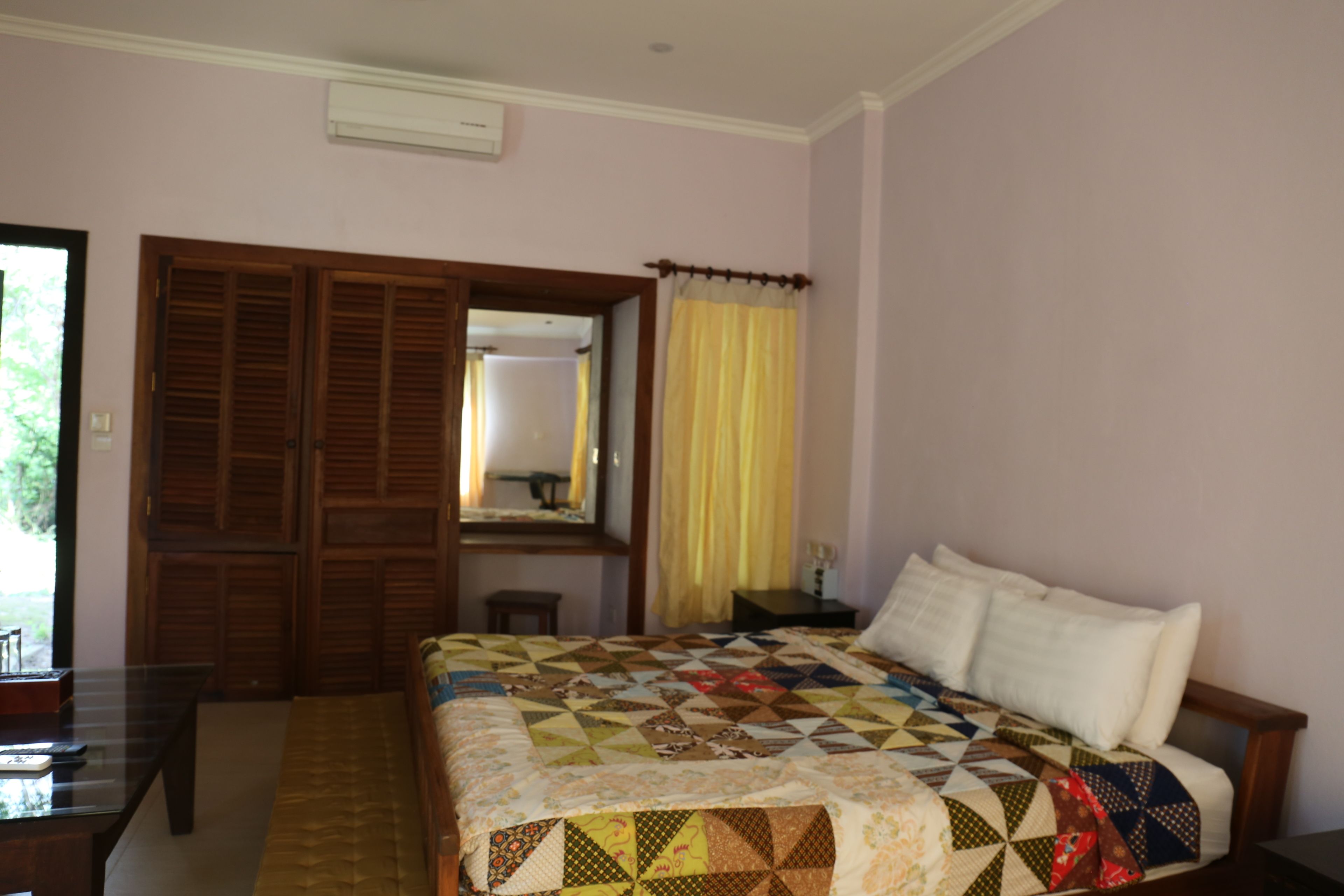 A bedroom with a bed, a wooden wardrobe, a mirror, a nightstand, and a window with yellow curtains.