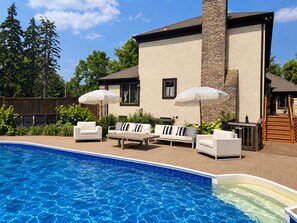 Una piscina con sillas de descanso y sombrillas, rodeada por una casa con fachada de ladrillo y una terraza de madera.