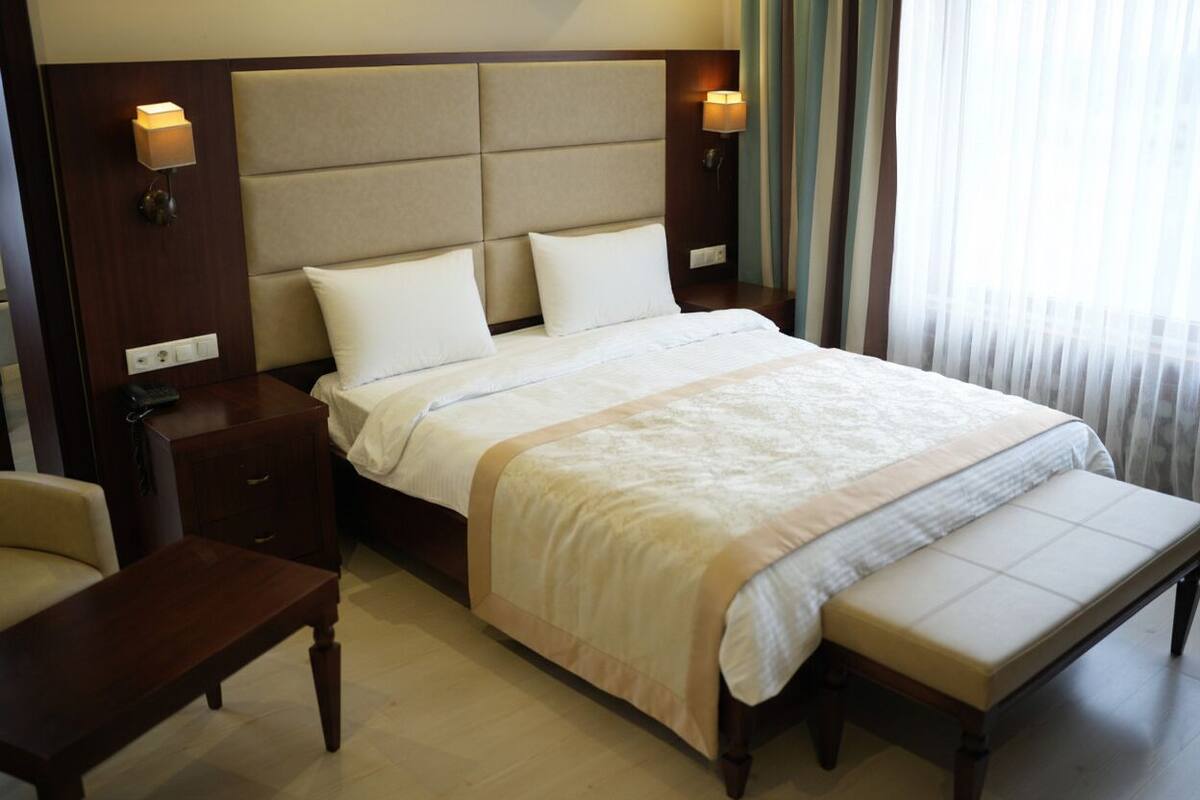 A hotel room with a large bed, a bedside table, a chair, and a bench.