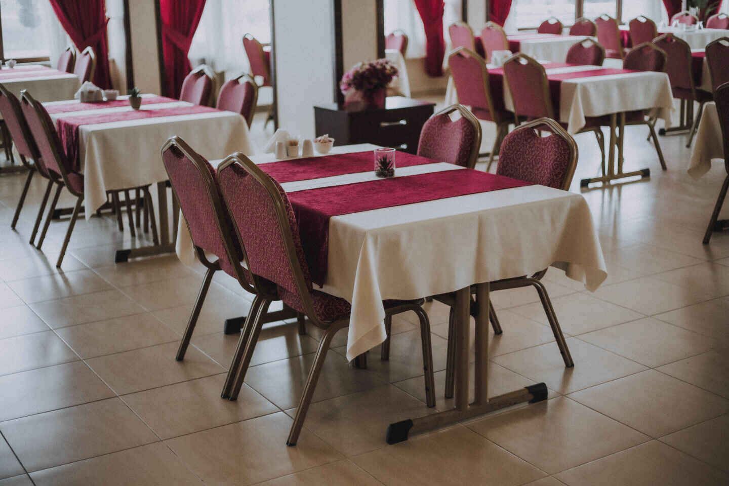 A dining area with tables covered in white tablecloths and red chair covers, arranged in rows.