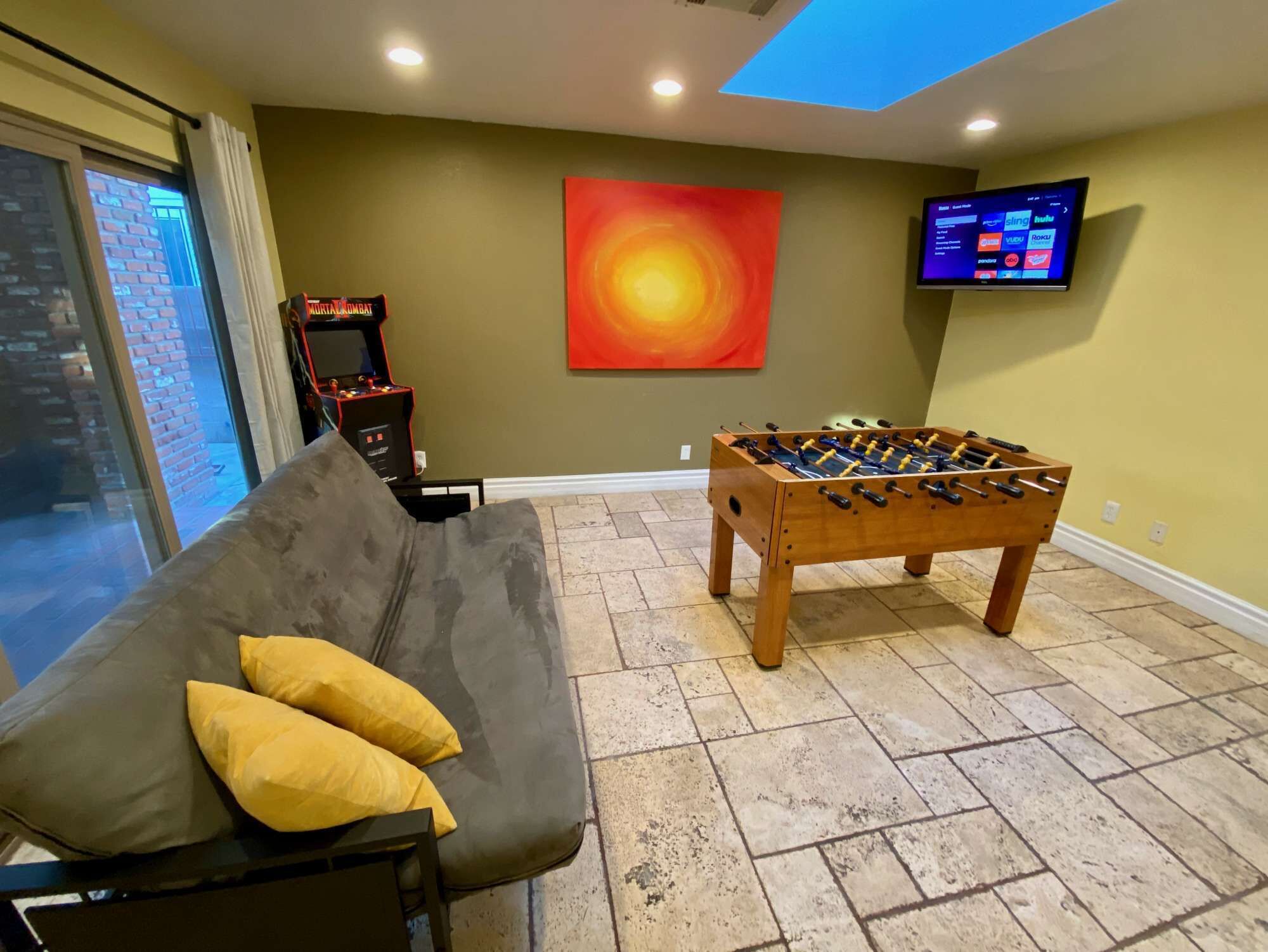 Una habitación con mesa de futbolín, sofá con cojines, televisor montado en la pared y un cuadro abstracto grande.
