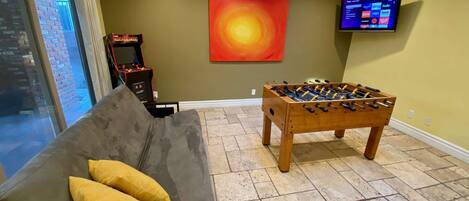 Una habitación con mesa de futbolín, sofá con cojines, televisor montado en la pared y un cuadro abstracto grande.