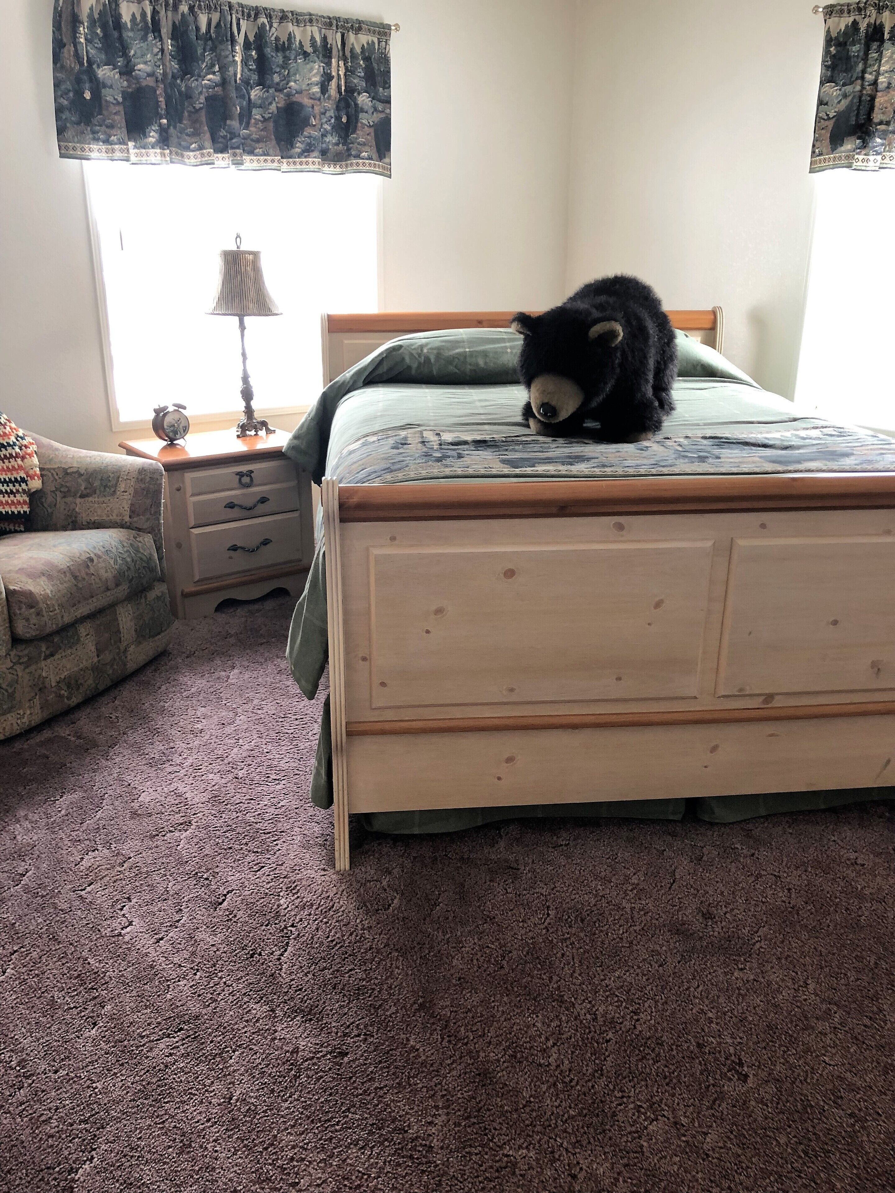 Un dormitorio con una cama de madera, una mesita de noche con lámpara, un sillón estampado y una ventana con cortinas.