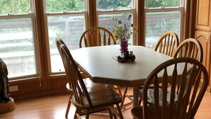 A dining room with a wooden table and chairs, a vase with flowers, and large windows with a view of greenery.