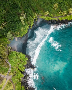 Une vue aérienne d’une zone côtière, avec un chemin dégagé menant à une plage, le tout bordé d’une végétation luxuriante et d’une côte rocheuse.