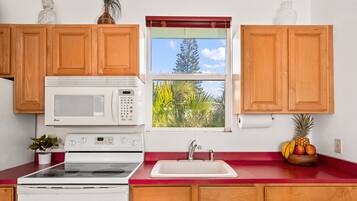 A kitchen with wooden cabinets, a white microwave, a stove, a sink, and a window with a view of trees and sky.