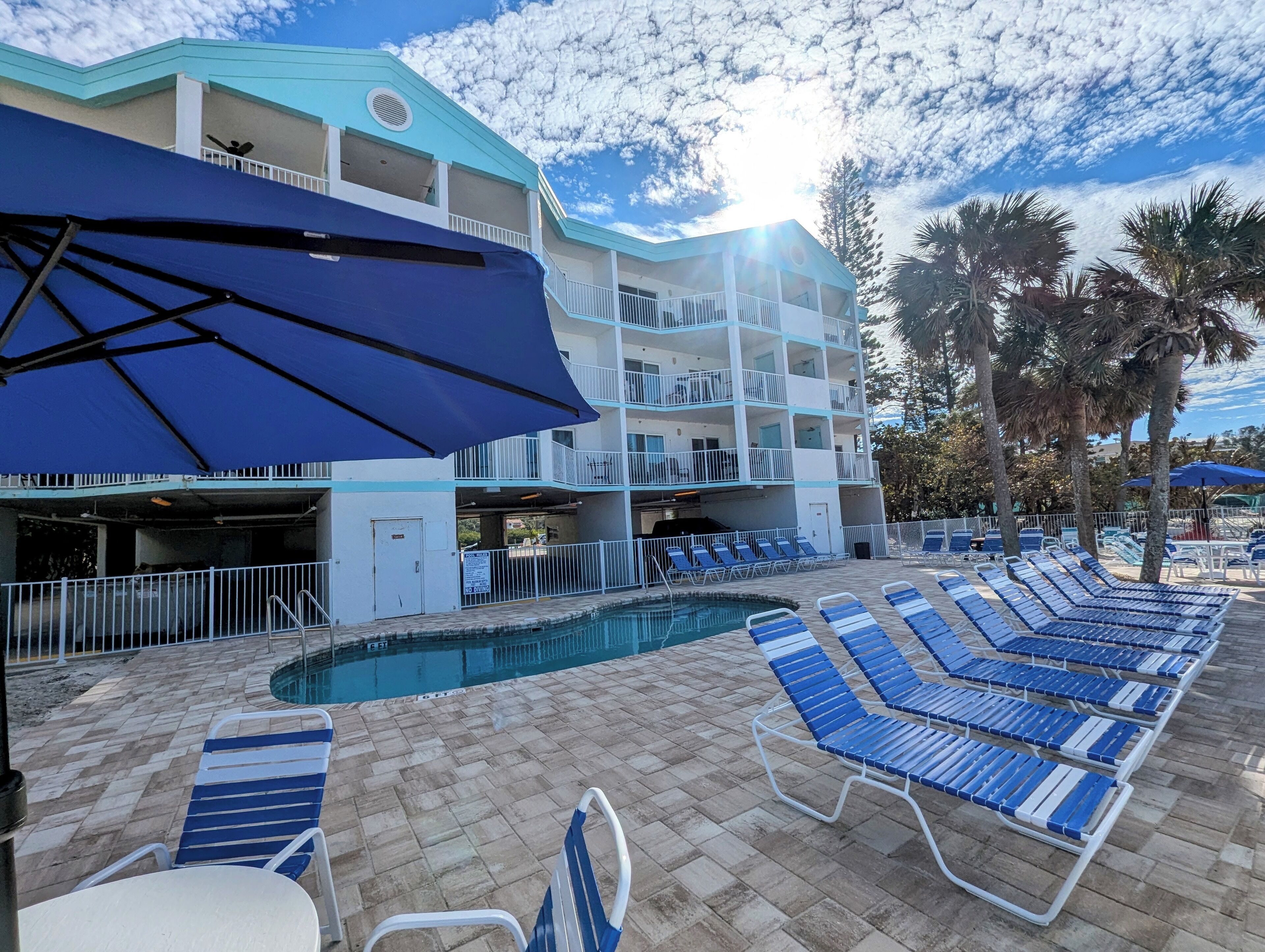 A hotel pool area with blue lounge chairs, a swimming pool, and a multi-story building with balconies.