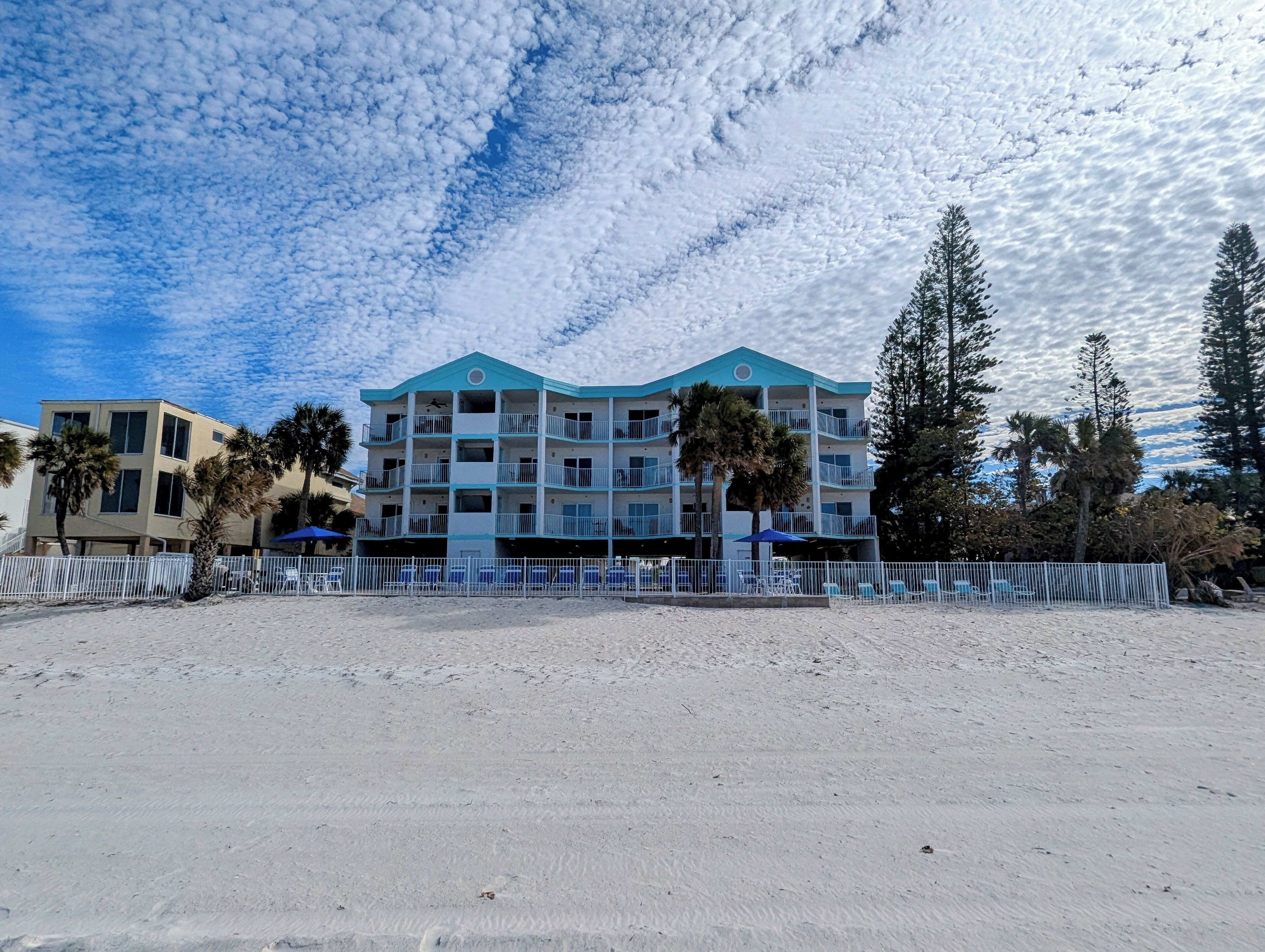 Ein Strandhotel mit mehreren Balkonen, ein sandiger Strand im Vordergrund und Palmen.